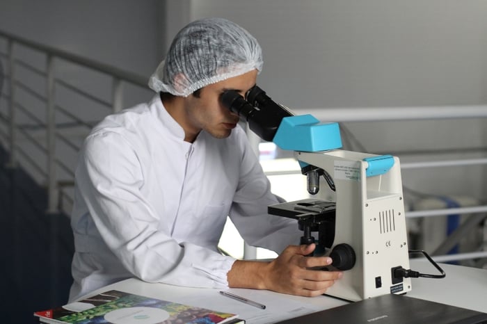 male doctor looking in a electronic microscope at a table with a pen paper magazines and laptop MedTech medical technology