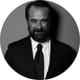 black and white photo, man standing by the wall, smiling, beard, white shirt, black suit, brown hair, black tie