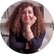 woman, having hand under her face, curly brown long hair, black tshirt, bracelet, necklace, shelves full of books behind her