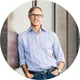 man, grey hair, wearing glasses, light blue shirt, jeans, he has hands in his pockets, standing next to the wall