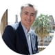 man smiling, standng, grey hair, glasses, light blue shirt, black suite, palm tree behind him, view on the building